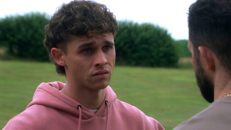emmerdale jacob sugden young man in pink hoodie looking concerned, standing outdoors.