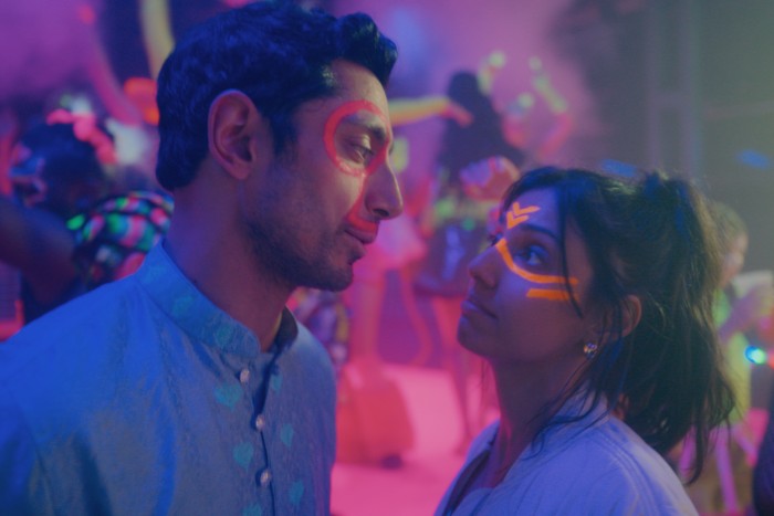 BAIT_First Looks_S1_Courtesy of AMAZON MGM STUDIOS (3) Riz Ahmed as Shah Latif and Ritu Arya as Yasmin in Bait. They are looking at each other in a neon-lit party, with facepaint on.