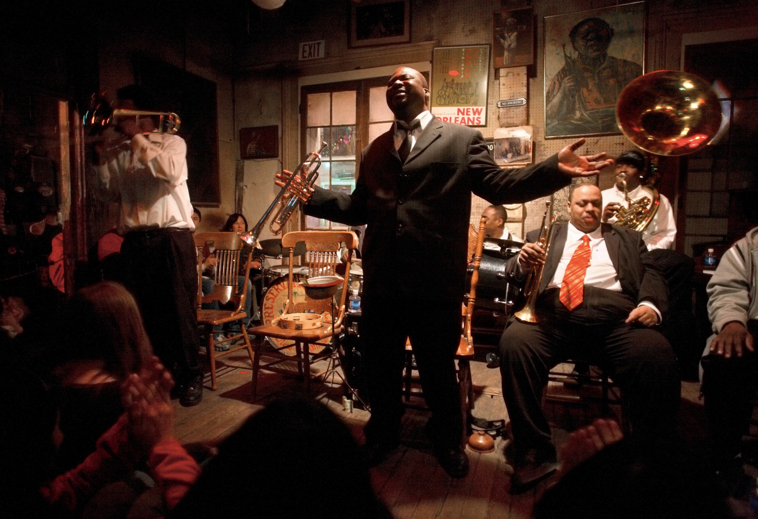 A singer in a dark suit raises his arms mid-performance inside a dimly lit New Orleans jazz club, surrounded by a brass band playing trumpet and tuba.