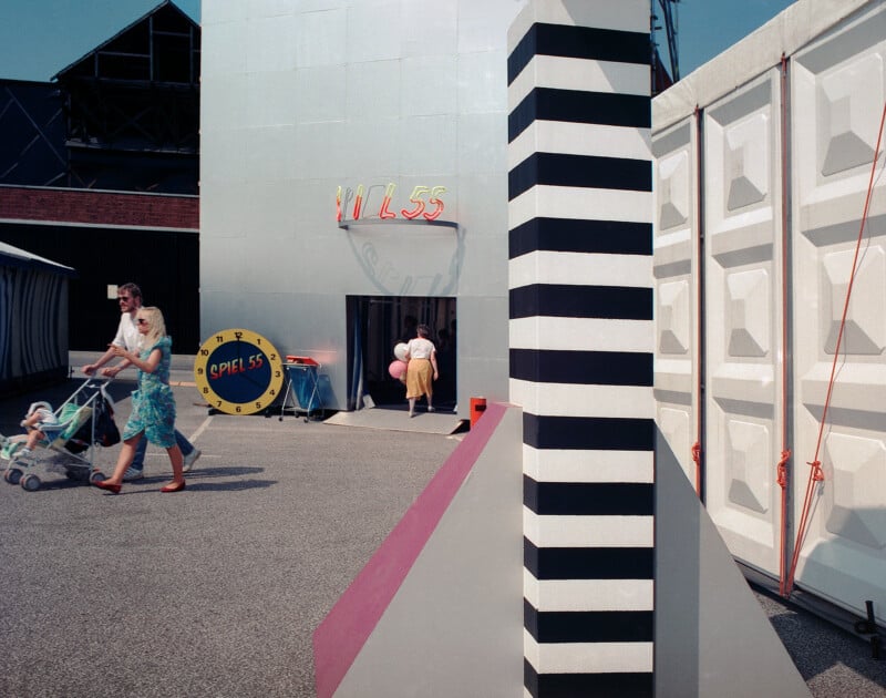 A family walks past a silver building marked "SPIEL 55" with a large clock-shaped sign, next to a black-and-white striped pillar.