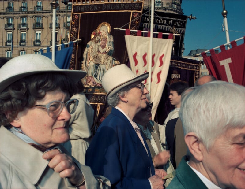 Several older women wearing hats and glasses stand in a crowd during a religious procession with banners depicting the Mater Dolorosa.