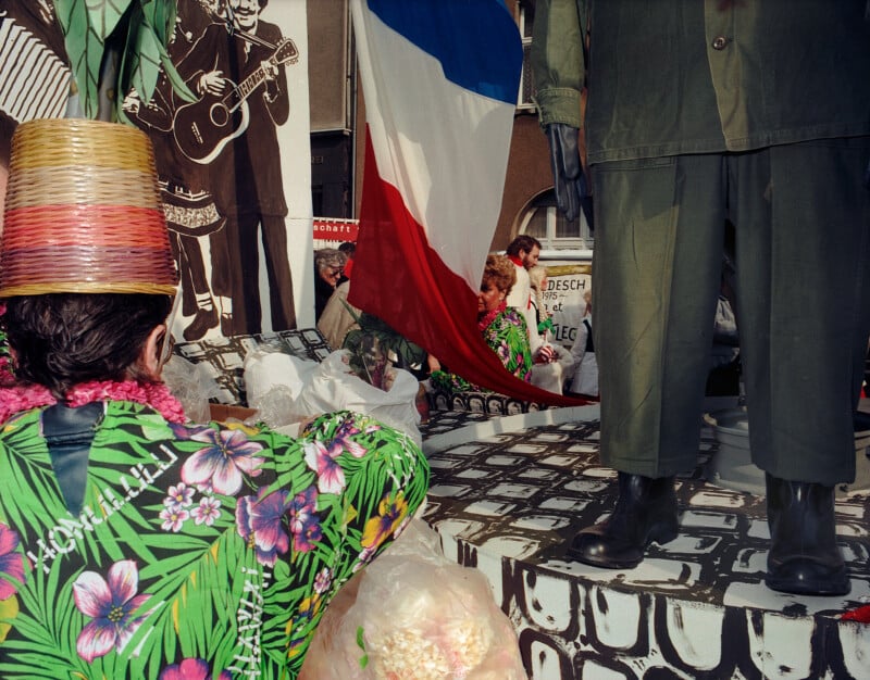 A person wearing a woven hat and a floral shirt stands next to a mannequin dressed in green military-style clothing and black boots, with a French flag draped nearby.