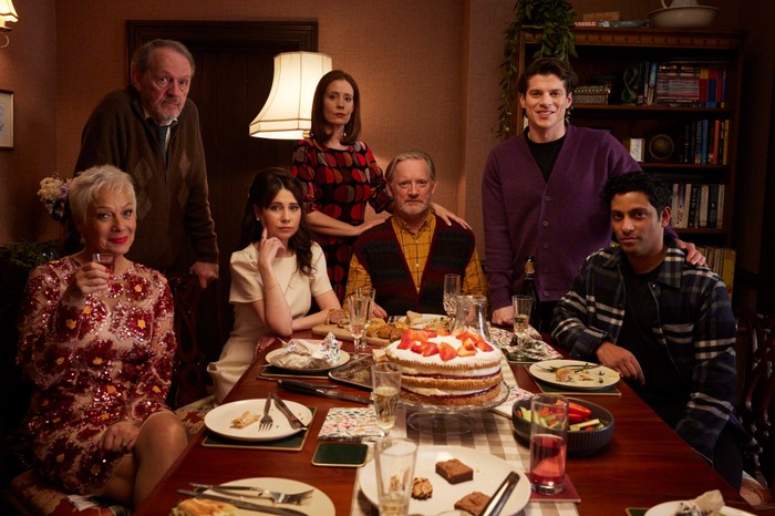 The cast of Forever Home, sat and stood around a dinner table, looking at the camera.