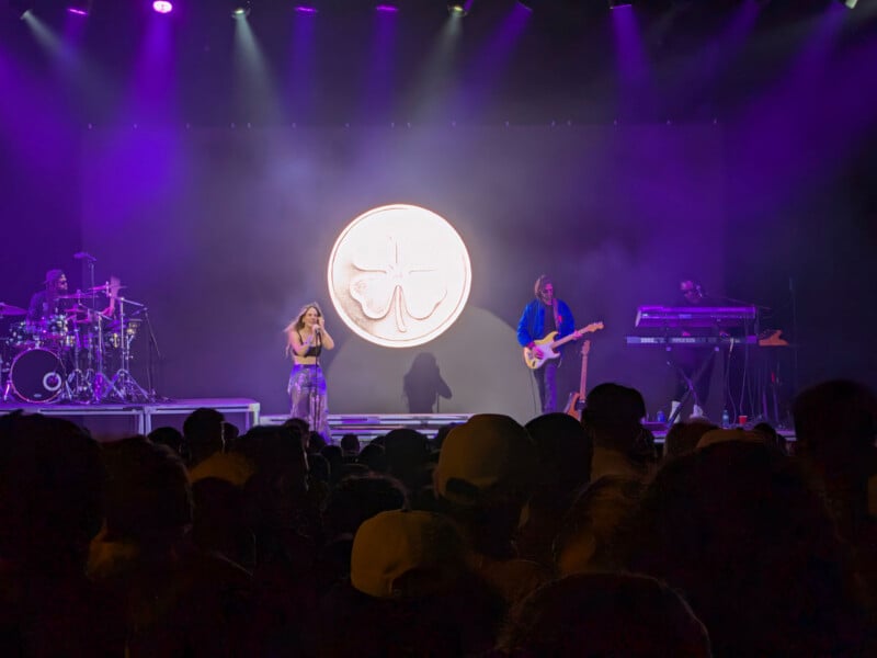 A band performs on a stage bathed in purple light, with a large, glowing four-leaf clover emblem projected on the backdrop behind them.