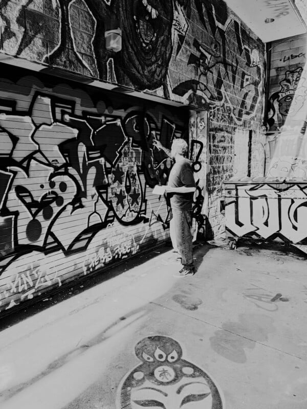 A person stands in an alleyway, spray-painting graffiti onto a metal garage door covered in various artistic designs.