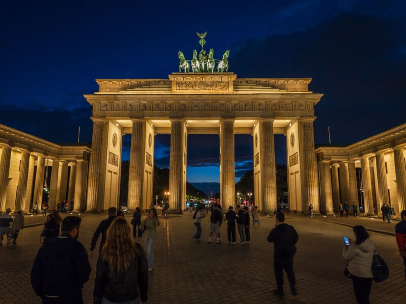 The Brandenburg Gate is illuminated at night, with people walking across the paved square in the foreground.