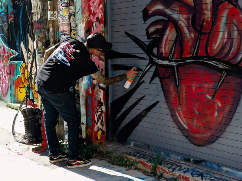 A person with tattoos on their arms wears a black t-shirt and cap while spray-painting a large red anatomical heart onto a grey metal garage door.