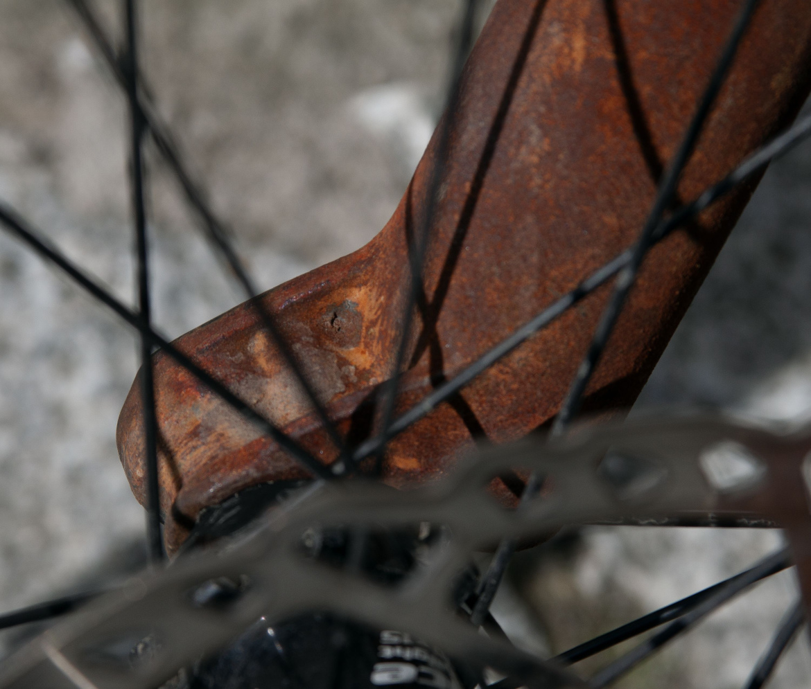 Close-up of a rusty bicycle part, showcasing the corroded metal and intricate details of the spokes and brake rotor in the background.
