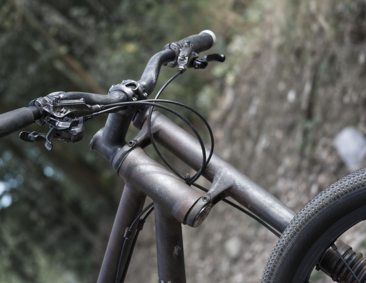Close-up view of a bicycle's handlebars and frame, featuring black grips, brake and gear levers, and a sleek, modern design. The background is blurred, suggesting an outdoor setting with greenery and rocky terrain.