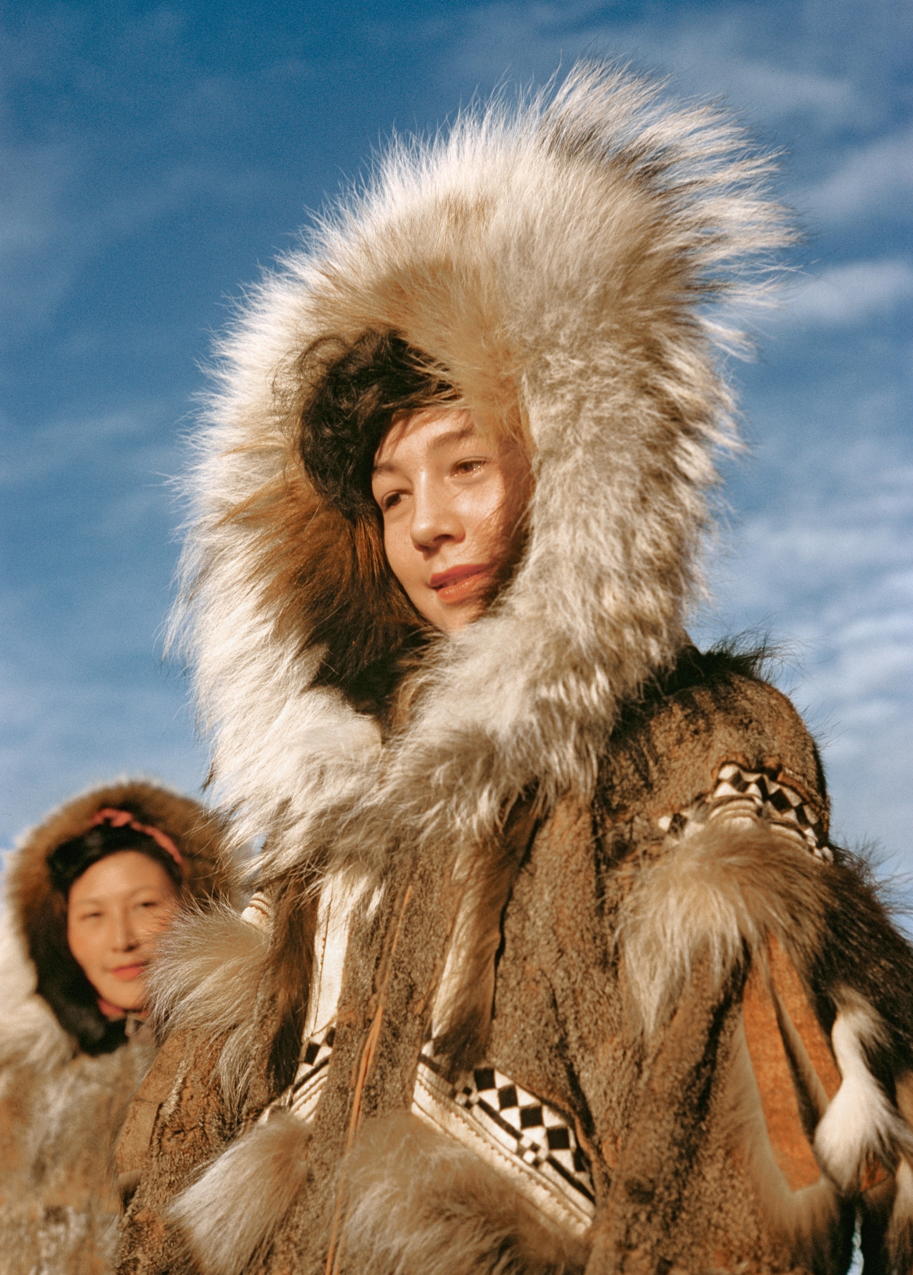 A young woman wears a thick fur-trimmed parka hood at an Alaskan Fourth of July celebration, with another similarly dressed woman standing behind her.