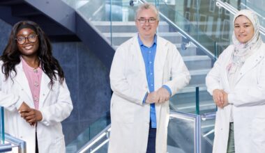 Two women and a man - diverse in age and ethnicity - pose in lab coats and smile in front of a metal and glass staircase.