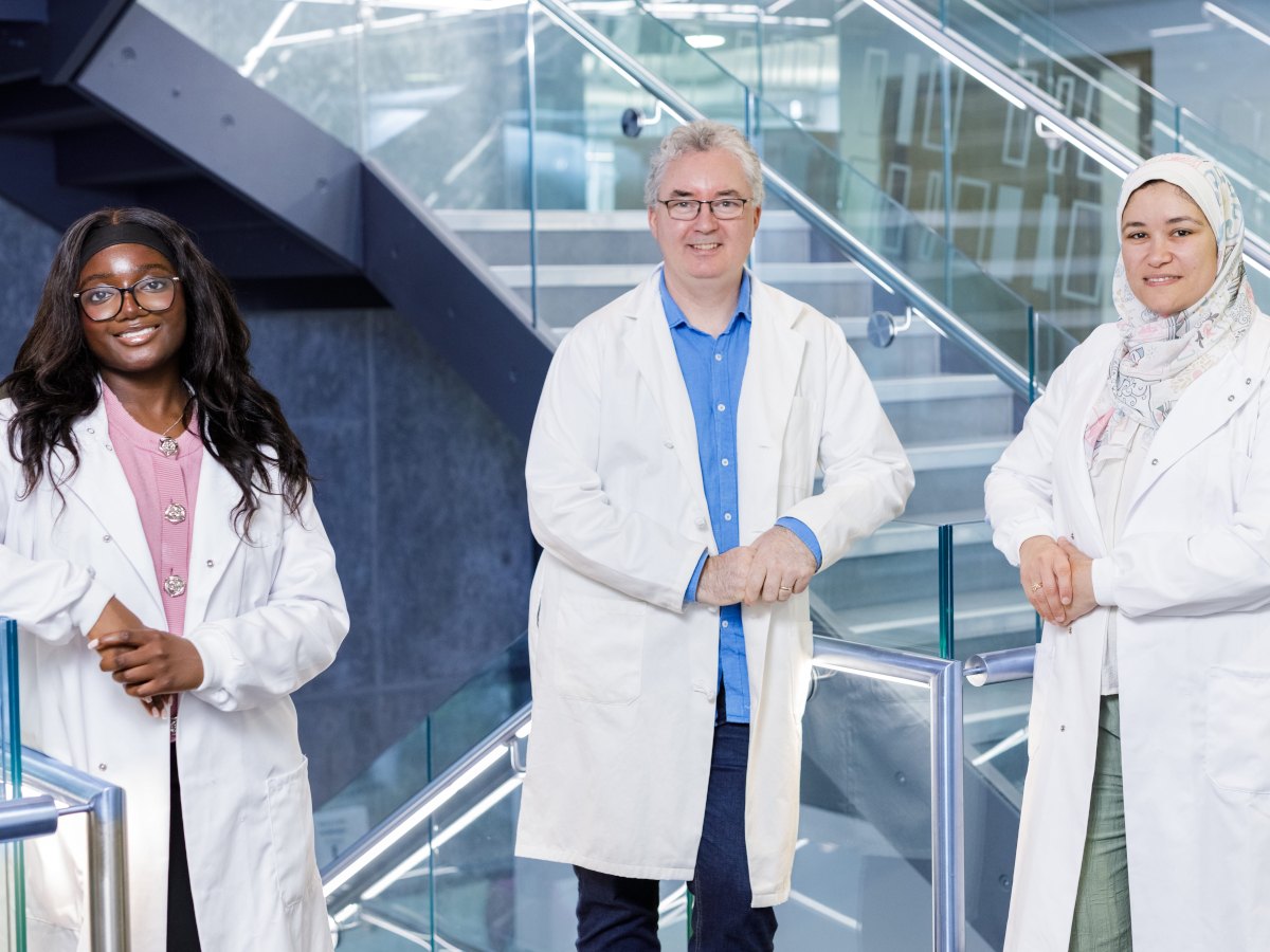Two women and a man - diverse in age and ethnicity - pose in lab coats and smile in front of a metal and glass staircase.