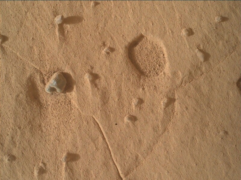 A close-up view of the dusty, orange-hued Martian surface featuring scattered small rocks, pebbles, and faint tracks.