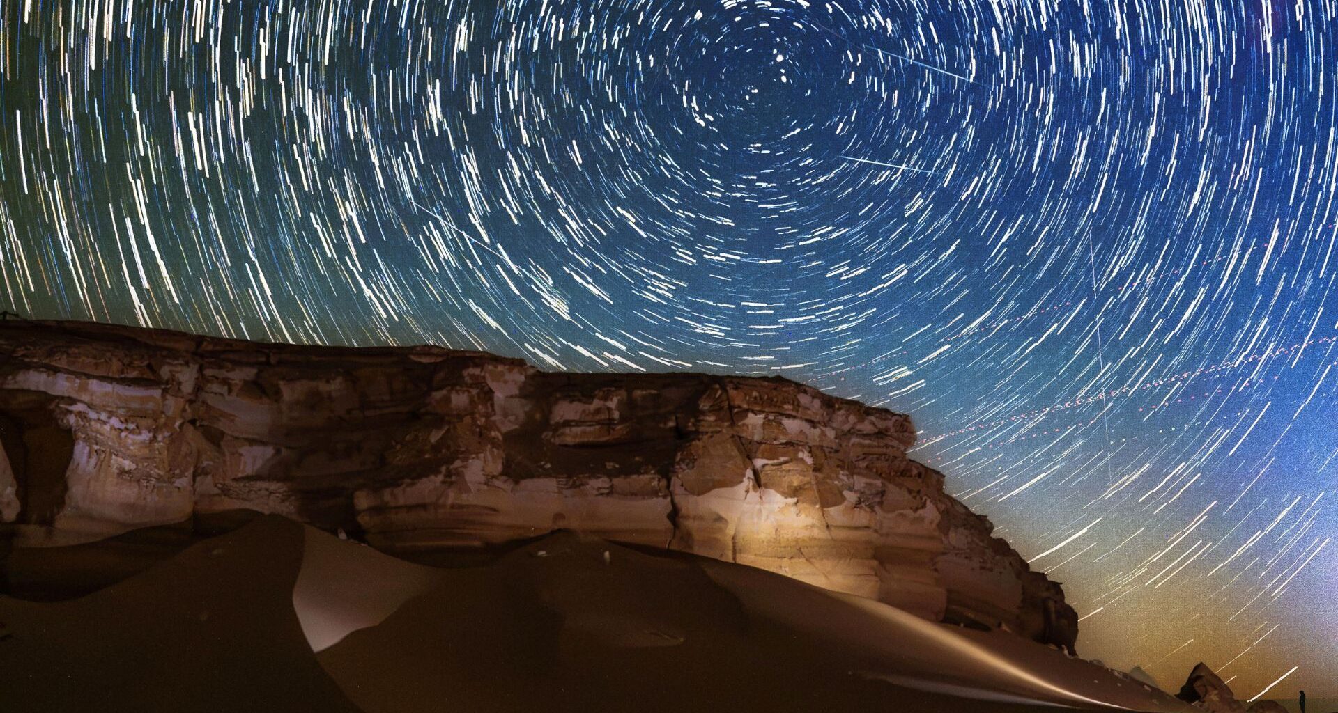 Star trails circle above a desert at night as meteors streak through the sky.