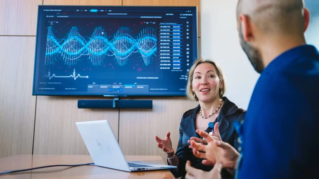 Physician speaking to a colleague in a conference room with a graph displayed on the screen behind her.