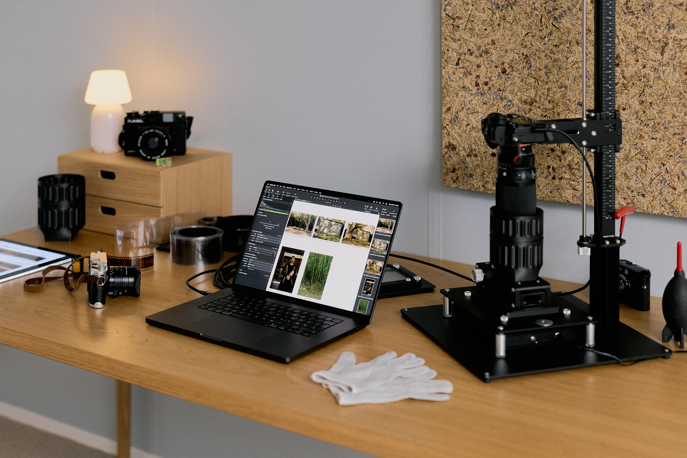 Workspace with a laptop displaying photo editing software, cameras, camera repair tools, and white gloves on a wooden desk