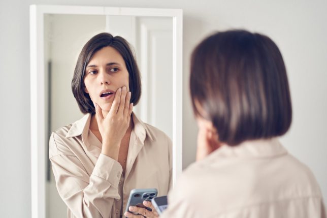 A young woman looks into a bedroom mirror, touching her cheek and checking her skin for blemishes while holding a smartphone, conveying concern, self-reflection and personal care moments.