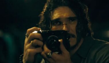 A man with long, dark, wavy hair and a mustache holds a black camera up to his face to take a photograph.