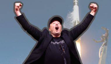 A triumphant Elon Musk, wearing a black baseball cap and a black t-shirt with 'FOUNDATION' written on it, stands with his arms raised high and mouth open in an excited expression. Behind him, a tall, slender white rocket is launching, emitting a bright orange flame and smoke from its base against a clear blue sky. A tall metal structure with robotic arms is visible to the right.