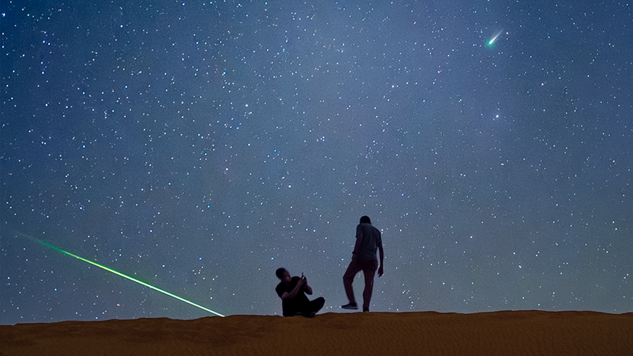 Two stargazers are silhouetted standing on a sand dune beneath a starry sky. A comet is visible to the upper right and a green meteorite can be seen streaking towards the ground to the left.