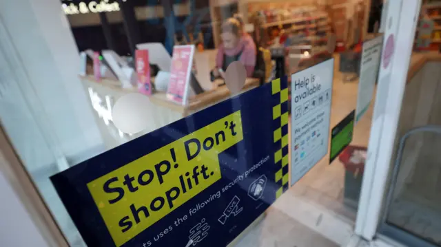 A sign in a shop window that says 'Stop! Don't shoplift'. In the background a woman can be seen at a counter, with shelves of good behind her