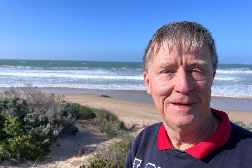 A man in a blue jumper and red colour in front of a beach with scrub