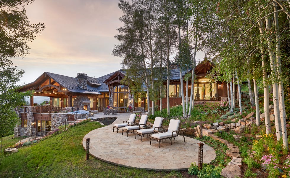 Modern mountain home surrounded by trees and landscaped garden, featuring a patio with lounge chairs and a hot tub.
