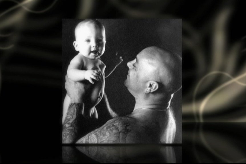 A black and white photo of a bald man with tattooed arms looking at a baby he's holding up while the baby looks at the camera.
