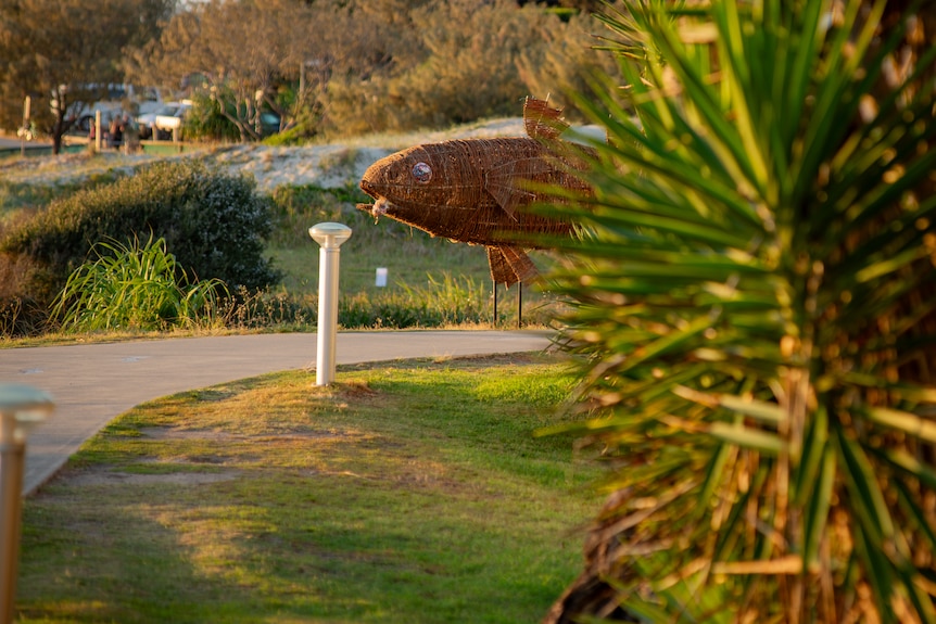 A woven sculpture of a fish alongside a concrete pathway on a beach foreshore.