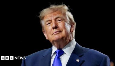 Donald Trump is wearing a navy suit jacket with a white shirt and a bright blue tie. He is smiling slightly and is standing in front of a black background.