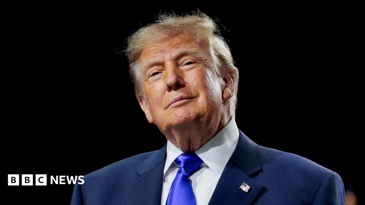 Donald Trump is wearing a navy suit jacket with a white shirt and a bright blue tie. He is smiling slightly and is standing in front of a black background.