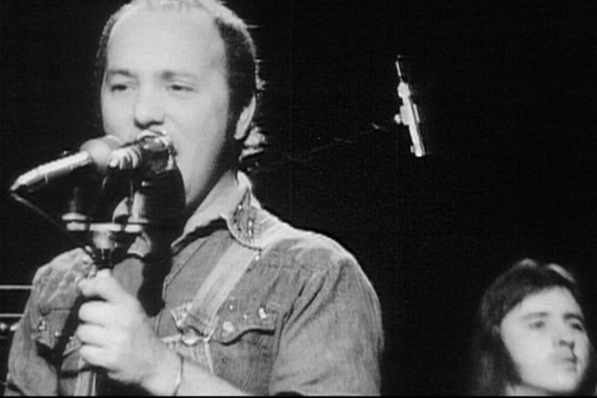 A black and white photo of a man in a denim jacke singing into a microphone with a long haired man seated behind him.