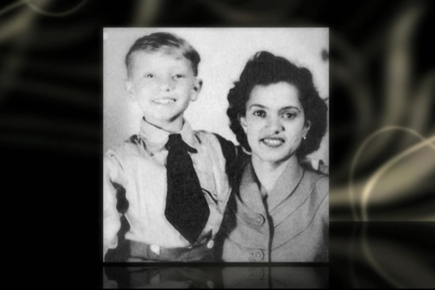 A black and white photo of a young boy in a collared shirt and tie standing with his arm around a woman in a dress.