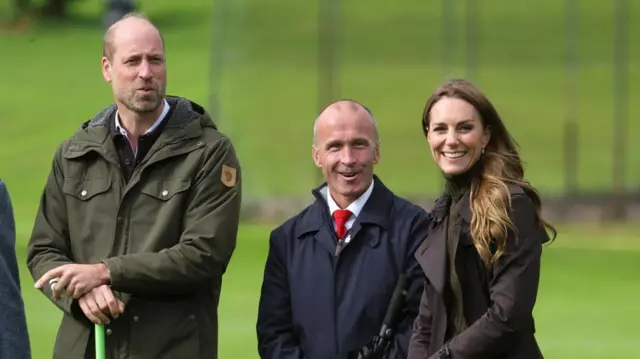 Craig Harrower, an older white man in a blue jacket, stands between the Prince and Princess of Wales surrounded by grass.