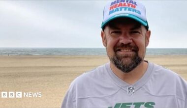 A man standing on a beach smiles at the camera in a head and shoulders image. He has a dark beard and is wearing a white baseball cap with blue peek. Across the front of the cap it says MENTAL HEALTH MATTERS in blue, red and turquoise letters. He is wearing a replica American football top in grey. It has JETS in green capitals across the chest.
