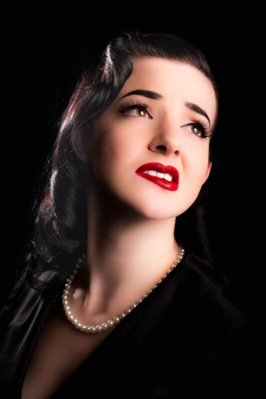 A woman with dark, vintage-style hair and red lipstick looks upward against a black background, wearing a pearl necklace and a black top.