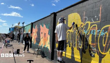 Artists paint murals on a large seaside wall. Some have ladders and stands. A man closest to the camera is spray painting yellow paint on to the black wall.