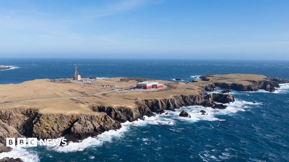 An aerial view of a long flat peninsula surrounded by dark blue sea. The coastline is rocky and the sky is blue. The island is brown and sparsely populated by several industrial buildings and a tall launch pad.
