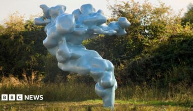 A large metallic green sculpture that is cloud-like rises out of the grass.