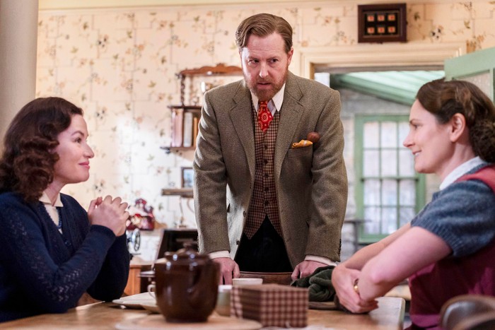 Three people in vintage clothing sit around a dining table indoors, while a bearded man stands between them and appears to be speaking.