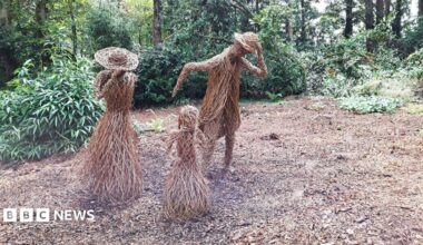 Willow sculptures depict a family of three leaning into the wind. The sculptures are life-sized. The two taller sculptures, a man and a woman, are looking down are are holding onto their hats. Trees and foliage is visible in the background. The area where they are stood is a relatively large space with no trees.
