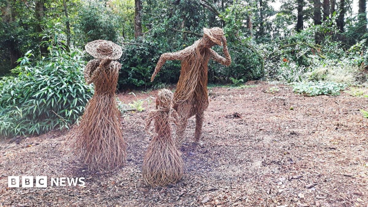 Willow sculptures depict a family of three leaning into the wind. The sculptures are life-sized. The two taller sculptures, a man and a woman, are looking down are are holding onto their hats. Trees and foliage is visible in the background. The area where they are stood is a relatively large space with no trees.