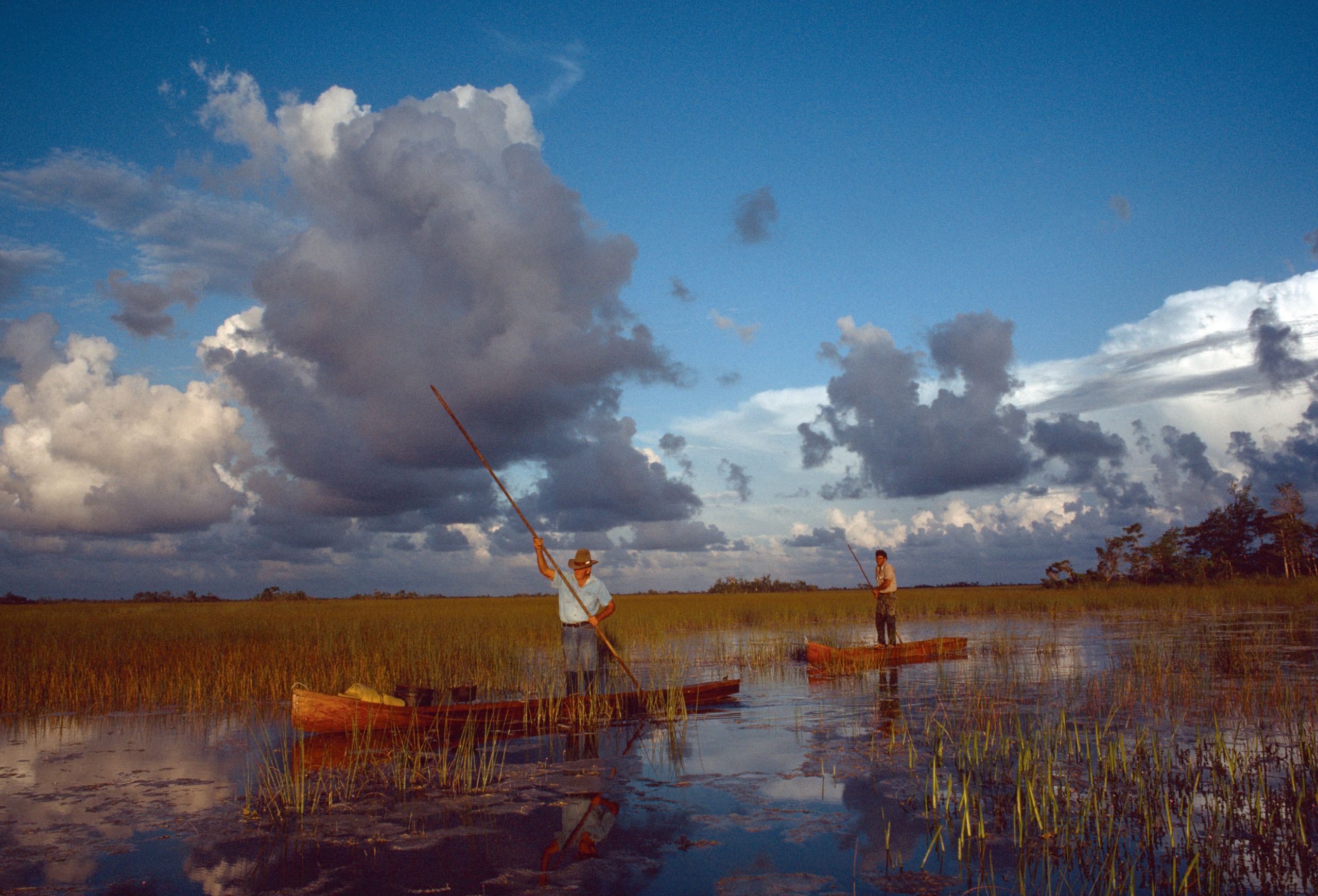Two men pole dugout canoes through the flooded sawgrass of the Florida Everglades beneath a dramatic sky of towering storm clouds.