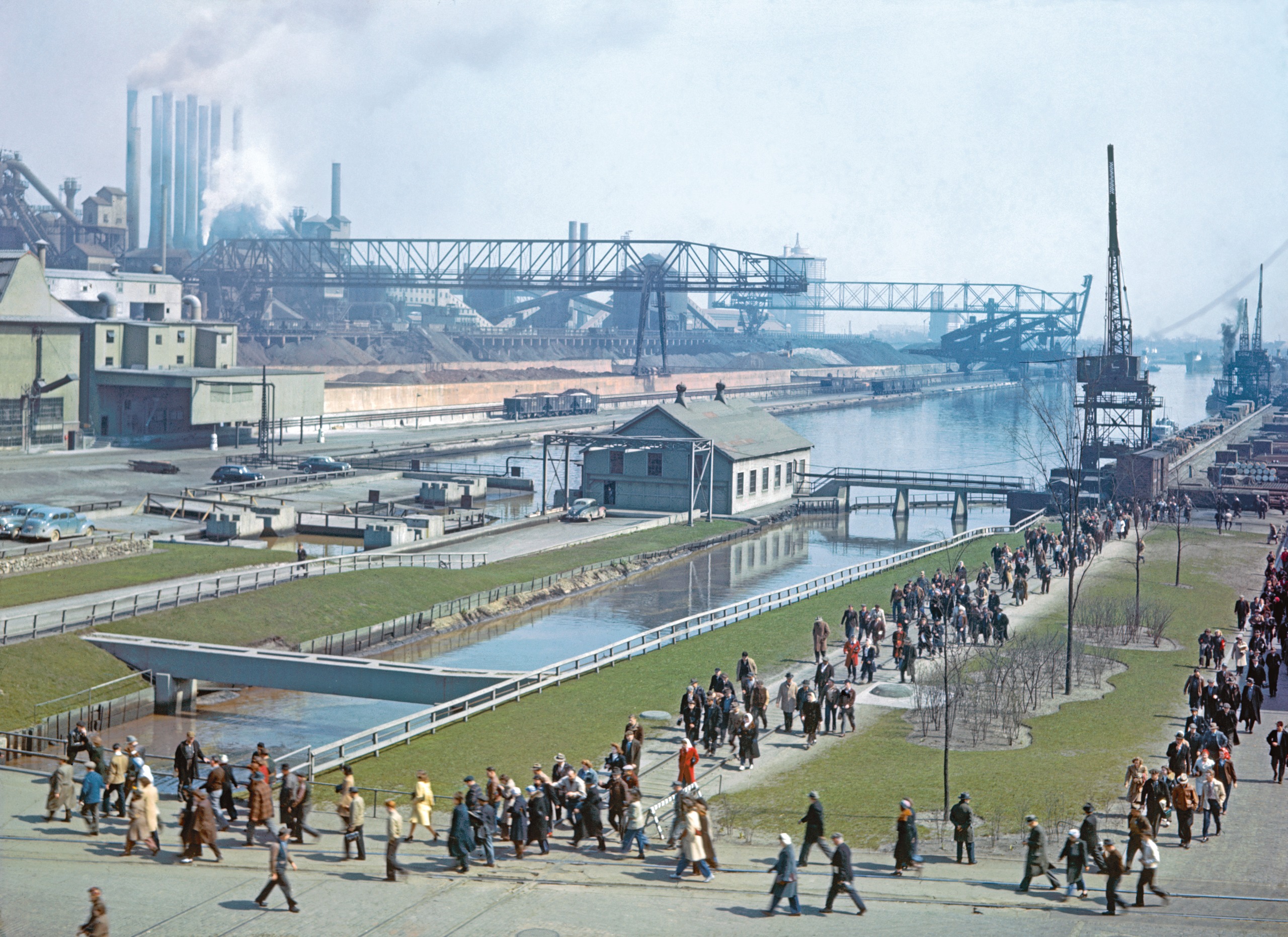 A crowd of factory workers walks along a riverside path past cranes, smokestacks and a steel plant belching smoke in the distance.