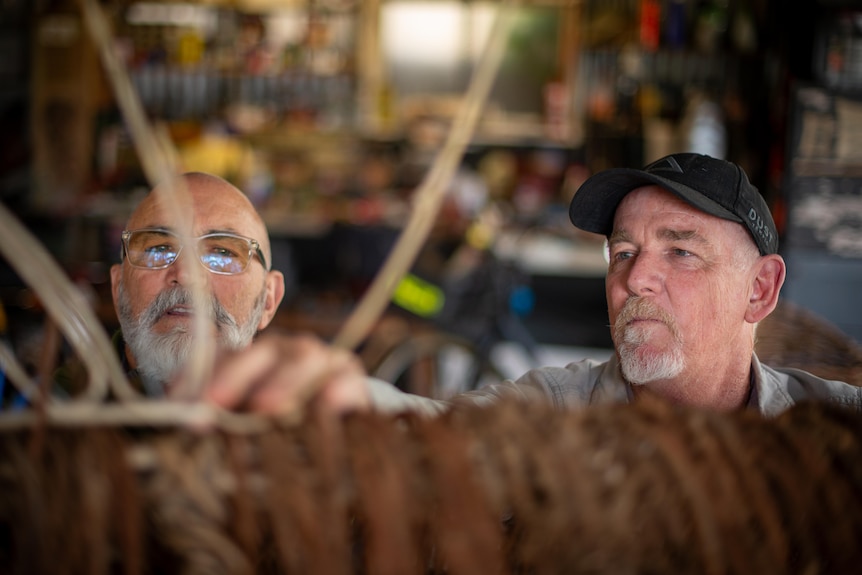 Two men peer out from behind a woven sculpture.