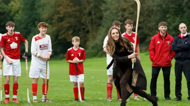 The Princess of Wales playing shinty on the pitch, holding a large stick and looking into distance as boys and men stand behind her.