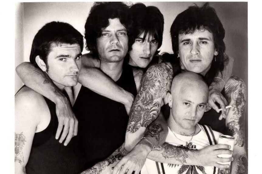 A black and white photo of four dark haired men and a bald man standing with their tattooed arms around each others' shoulders.