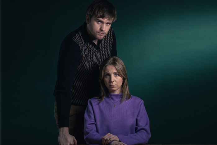 Emmerdale's Belle and Tom King In a stylised shot to promote a domestic abuse storyline on Emmerdale, Belle sits with a purple top on while Tom stands behind her menacingly. She has her hands clasped in front of her and a concerned look on her face. Tom is in black, with one arm in front, his hand clenched in a fist next to Belle. He has a stern look on his face. They are in front of a dark green background.