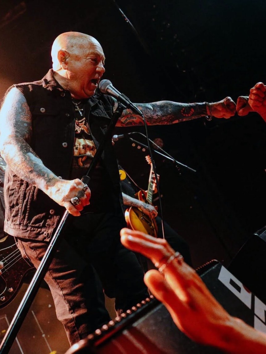 A bald man with tattooed arms in a black sleeveless shirt holds a microphone stand in hand and stretches the other arm out.