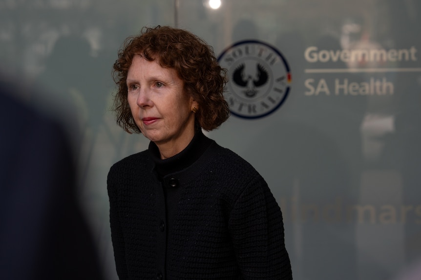 A woman with dark curling hair is standing in front of a glass building which has an SA Health logo.